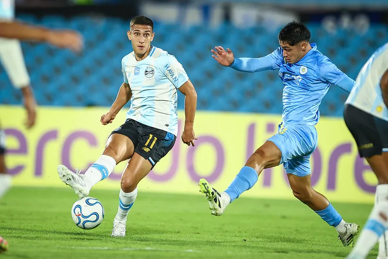 Jogadores de futebol em campo, com uniformes de Grêmio-RS e Deportivo Riestra, durante partida internacional.