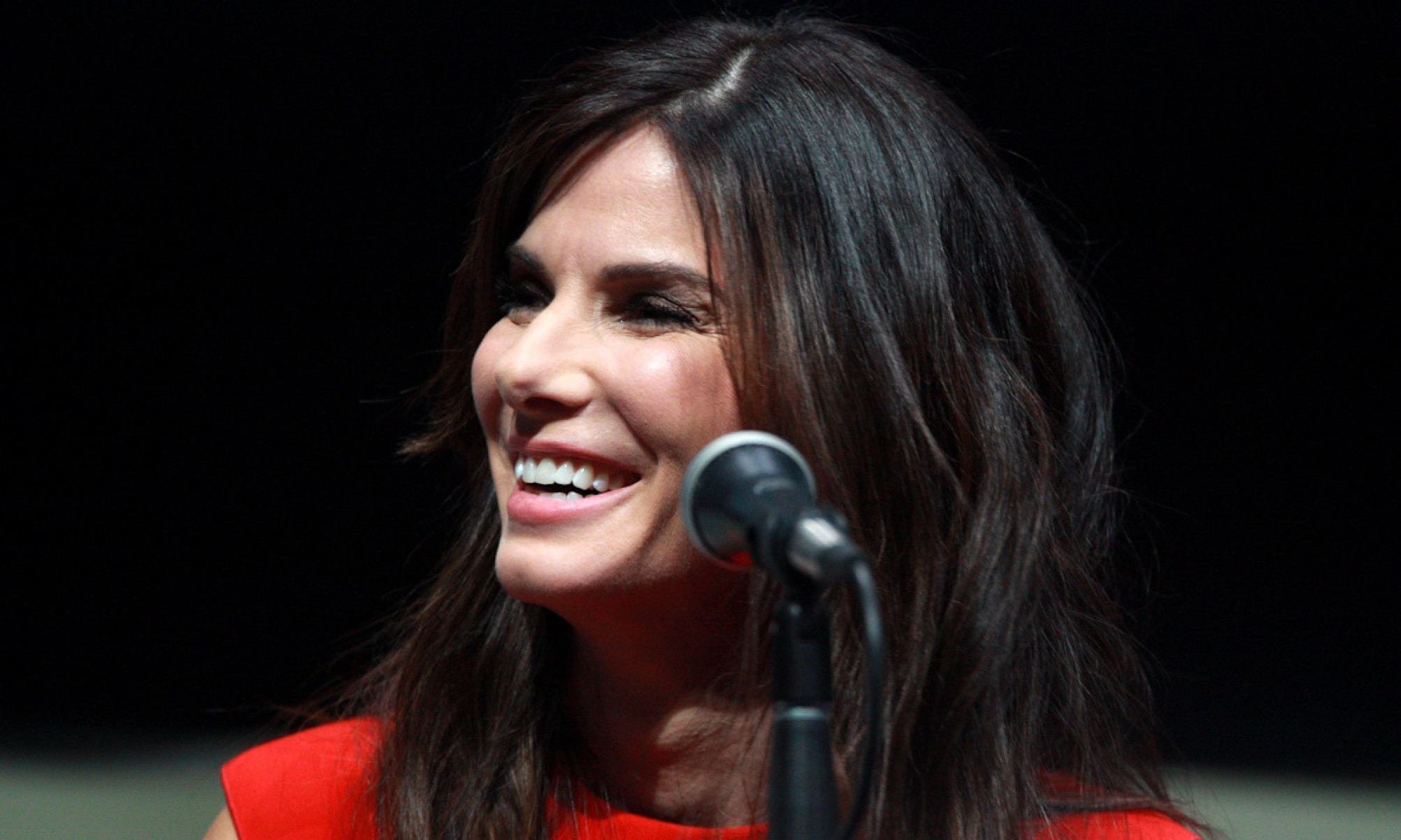 Sandra Bullock posa sorrindo em frente a tela de celular com notícias de milhões de seguidores em redes sociais.