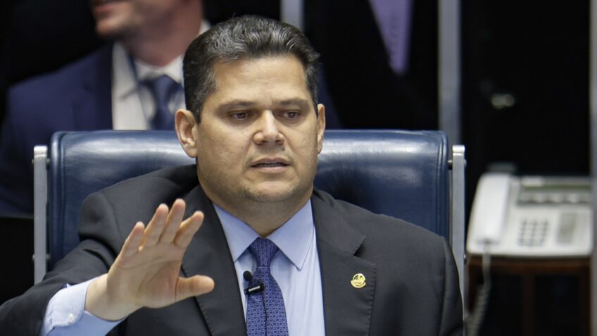 Político Alcolumbre sentado em frente a uma mesa de reunião, com um senador ao lado, ambos olhando para um papel com números.