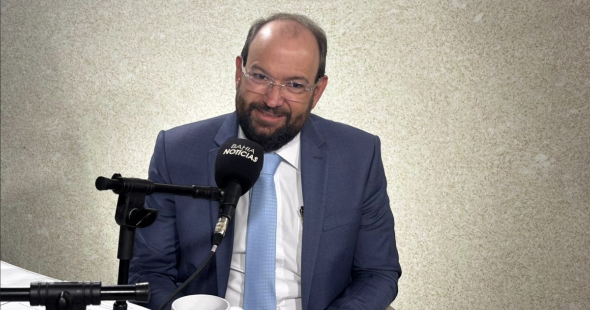 O presidente do TRE-BA é fotografado sorrindo, cercado por bandeiras brasileiras, em um cenário de auditoria lotada.