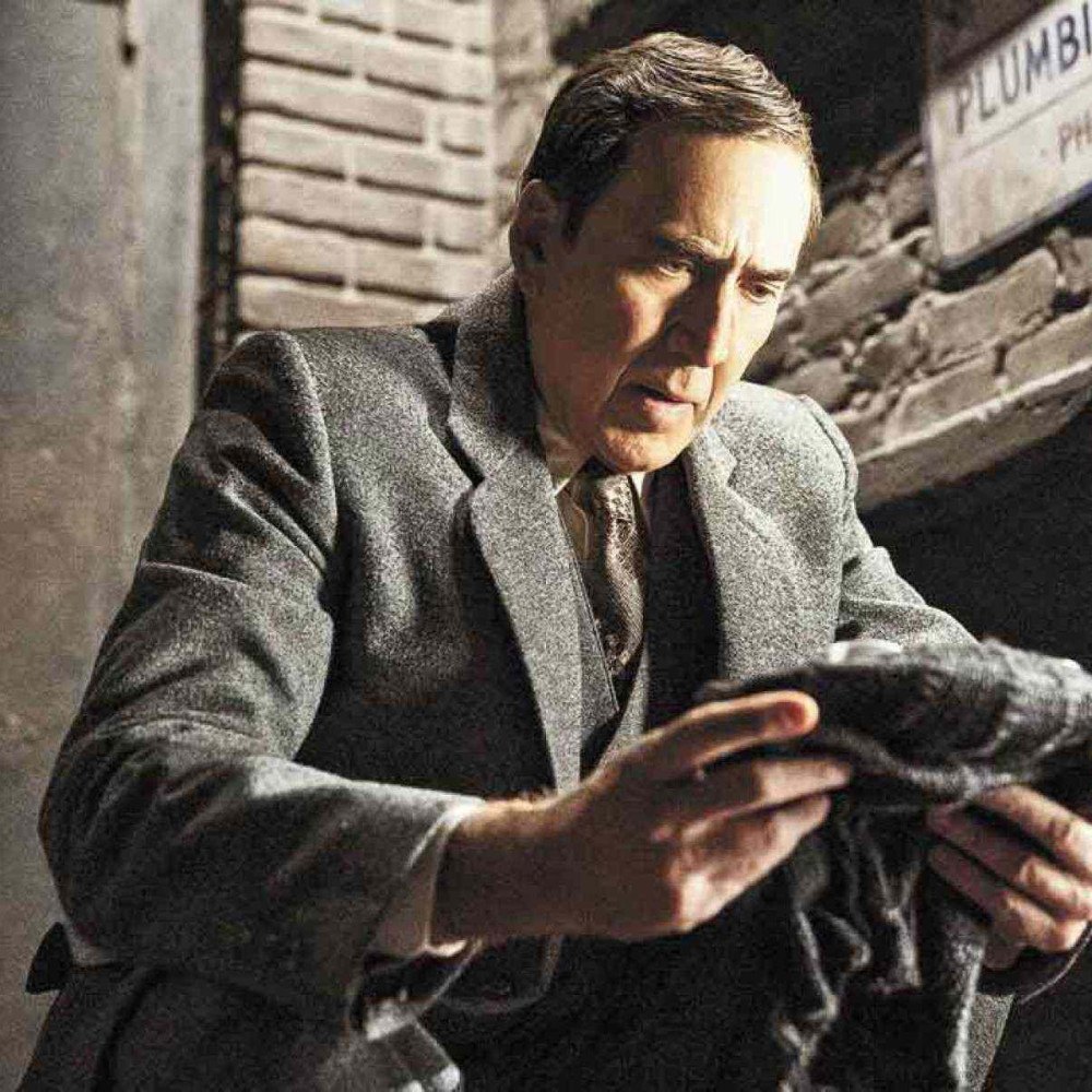 Nicolas Cage vestido como clone do Homem-Aranha, em cena de notícia jornalística, com fundo de cinema em ruínas.