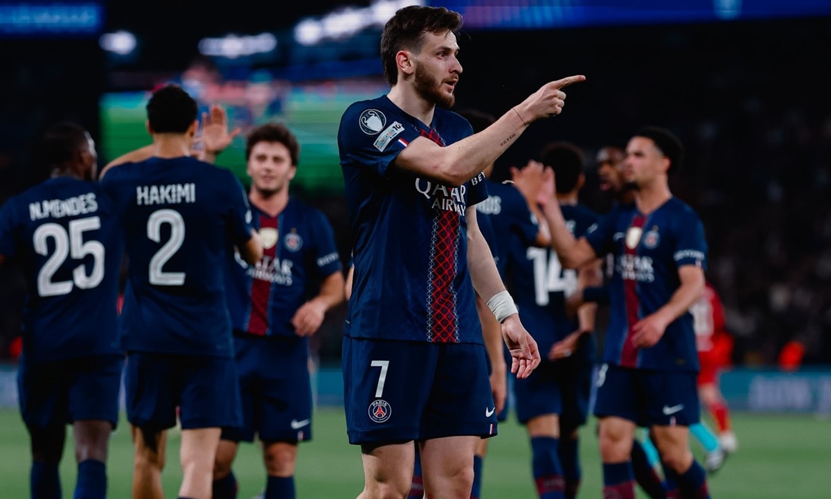 Jornalista sentado à mesa de trabalho com uma tela de computador e um jornal aberto, mostrando a notícia de Liverpool x PSG.