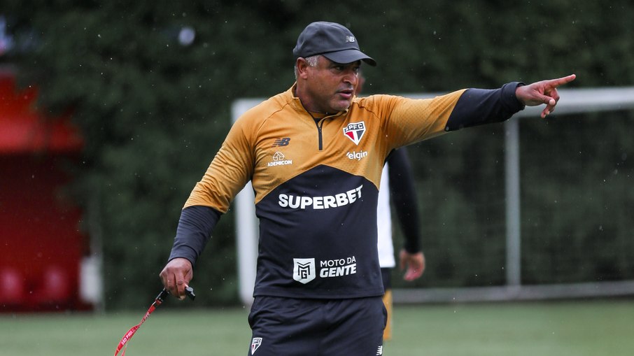 Jornalista sentado à frente de uma tela de TV exibindo um jogo de futebol entre São Paulo e Juventude.
