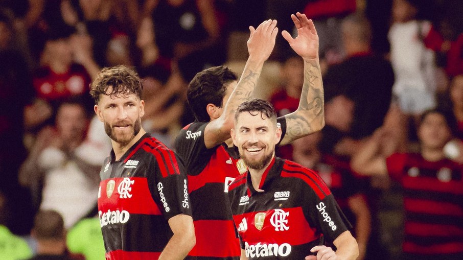 Jogadores de futebol em ação no estádio, com a bandeira do Flamengo ao fundo.