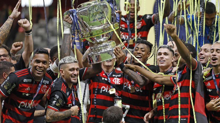 Jogadores de futebol em ação no estádio, com a bandeira do Flamengo ao fundo.