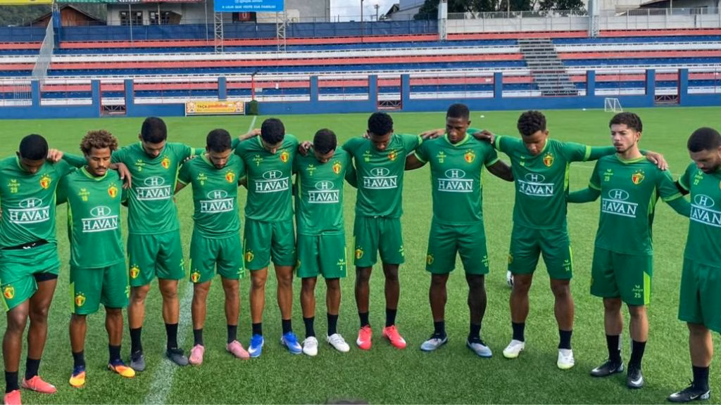 Time de Brusque em ação no campo de futebol, jogadores em movimento, torcida ao fundo.