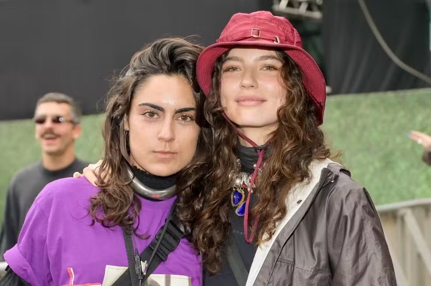 Atriz Alanis Guillen com expressão séria em frente a uma reportagem sobre medida protetiva contra ex-namorada.