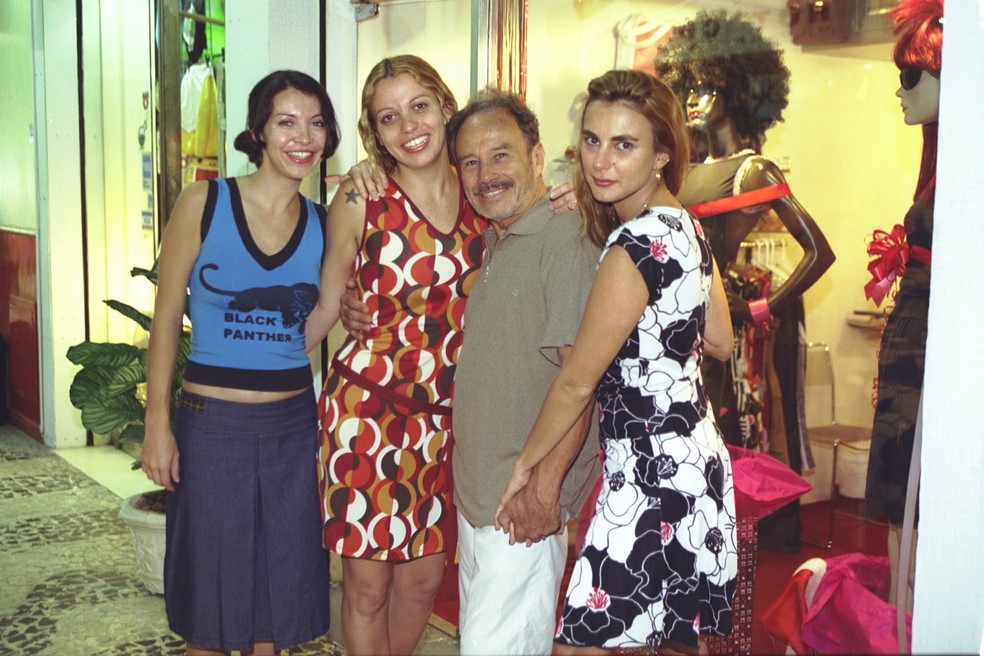 Mulher de sorriso discreto e vestido casual, sentada ao lado de um repórter, com um fundo de noticiário em segundo plano.