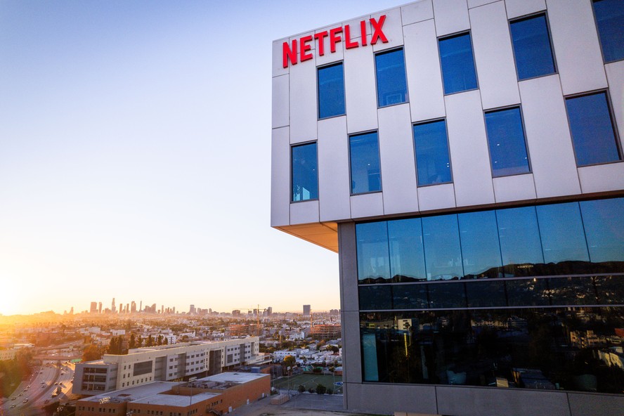 Imagem de um jornalista sentado à mesa, com um jornal aberto mostrando a notícia sobre a ação do Texas contra a Netflix.