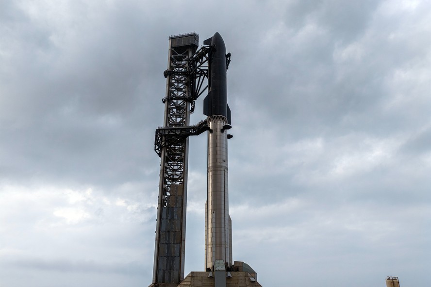 Imagem de uma equipe de jornalistas em volta de uma tela de TV mostrando a SpaceX Starship em lançamento.