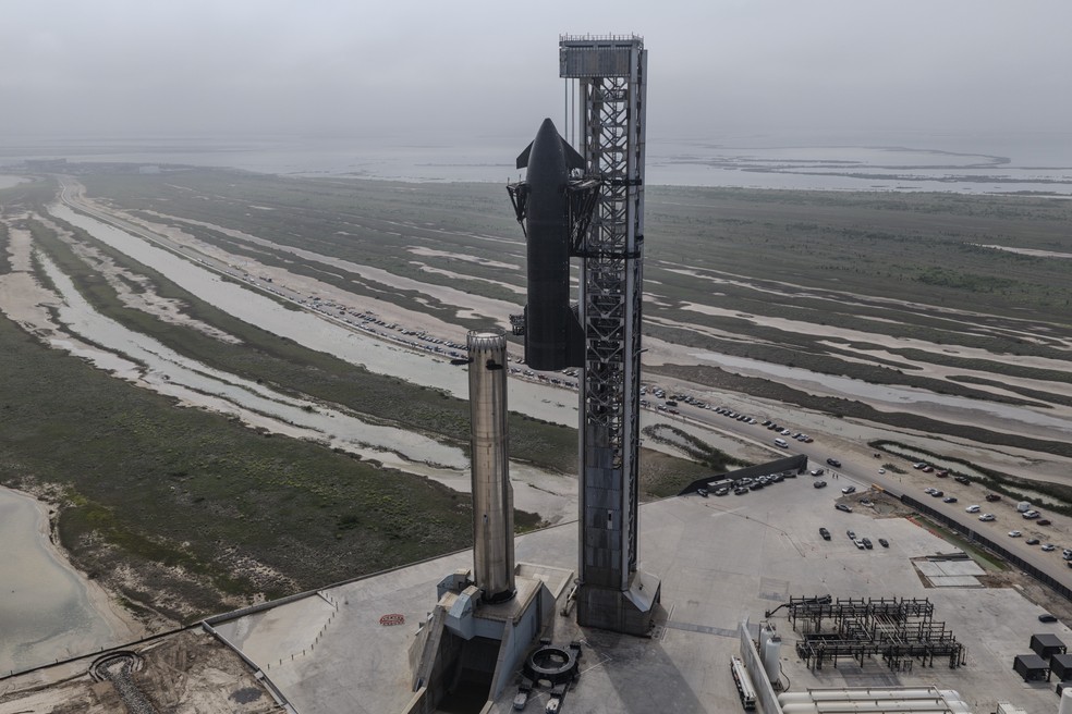 Imagem de uma equipe de jornalistas em volta de uma tela de TV mostrando a SpaceX Starship em lançamento.