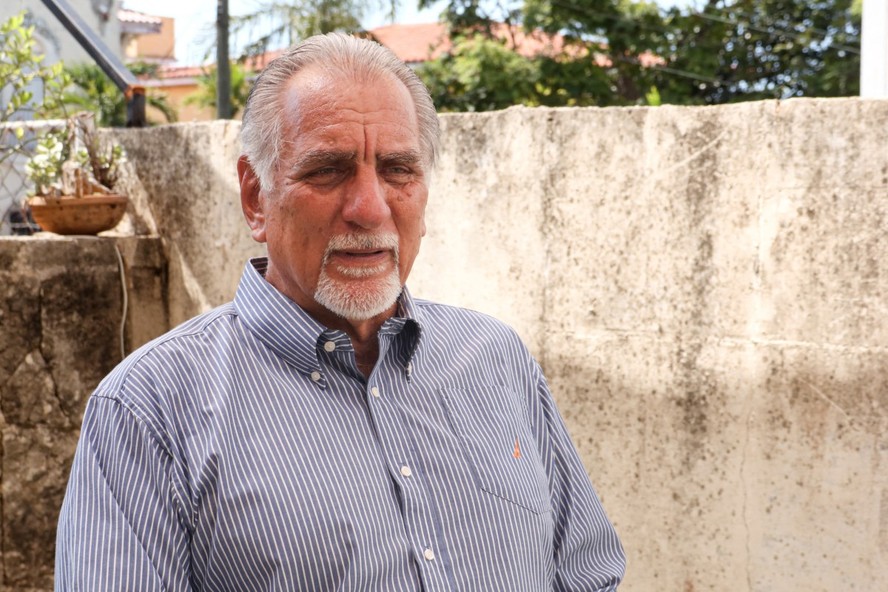 Homem de meia-idade com expressão séria, sentado em frente a uma mesa de madeira, com papéis e documentos espalhados ao redor.