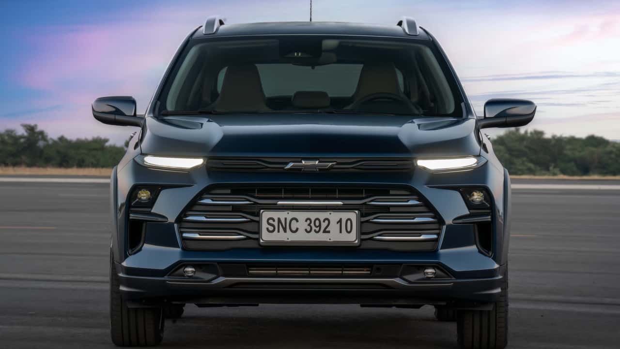 Jornalista segurando um exemplar do Chevrolet Sonic 2027 em frente a uma grande placa com a notícia de 14 mil unidades vendidas.