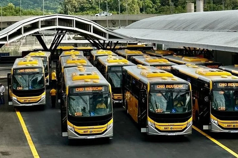 Imagem de uma linha de ônibus do BRT em movimento, com pessoas a bordo, passando por uma área urbanizada.