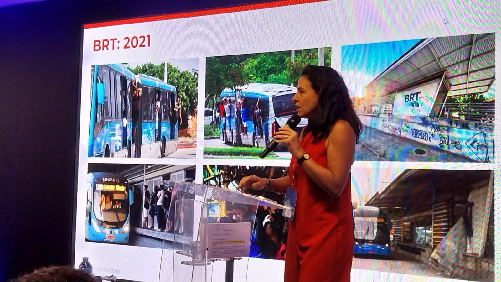 Imagem de uma linha de ônibus do BRT em movimento, com pessoas a bordo, passando por uma área urbanizada.
