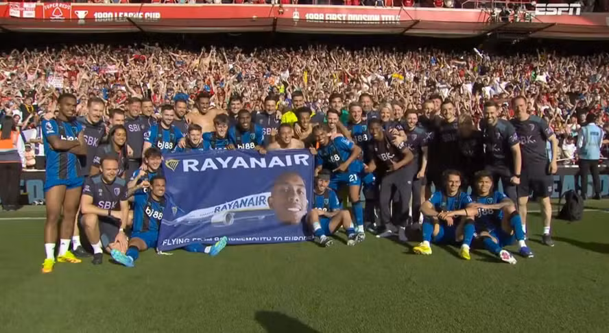 Jogadores do Bournemouth celebram classificação à Liga Europa erguendo bandeira com rosto de Rayan.
