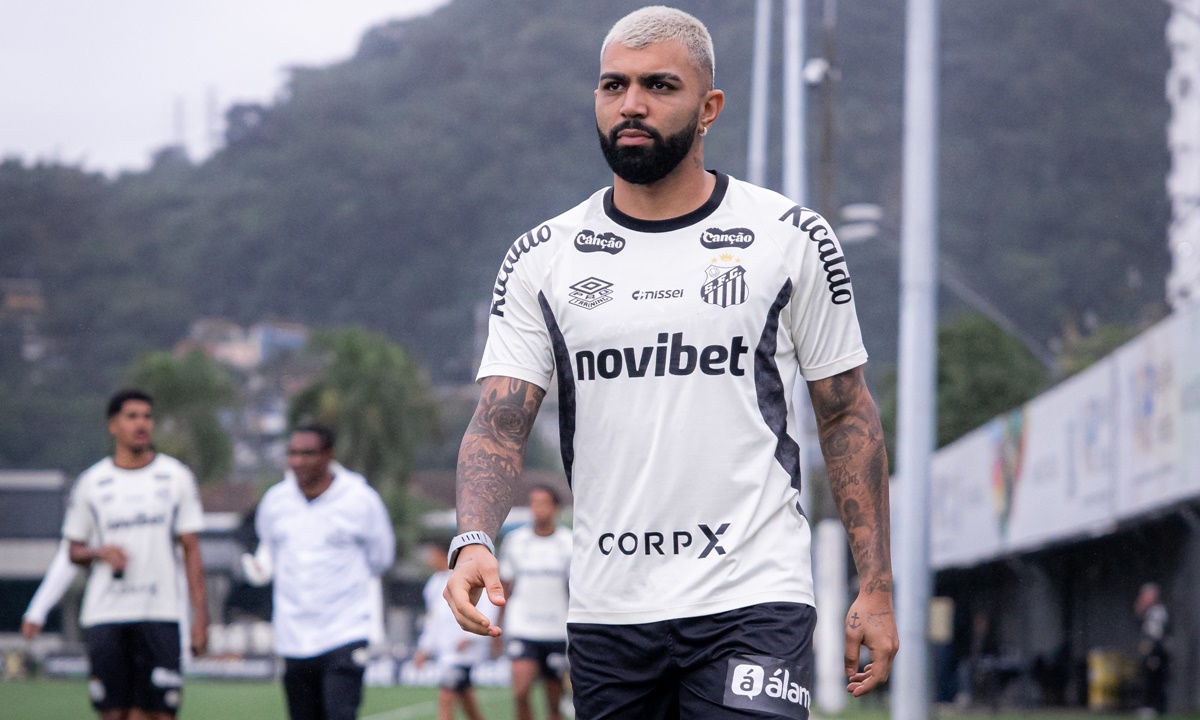 Jornalista sentado à mesa com computador e papel, revisando informações sobre partida de futebol.