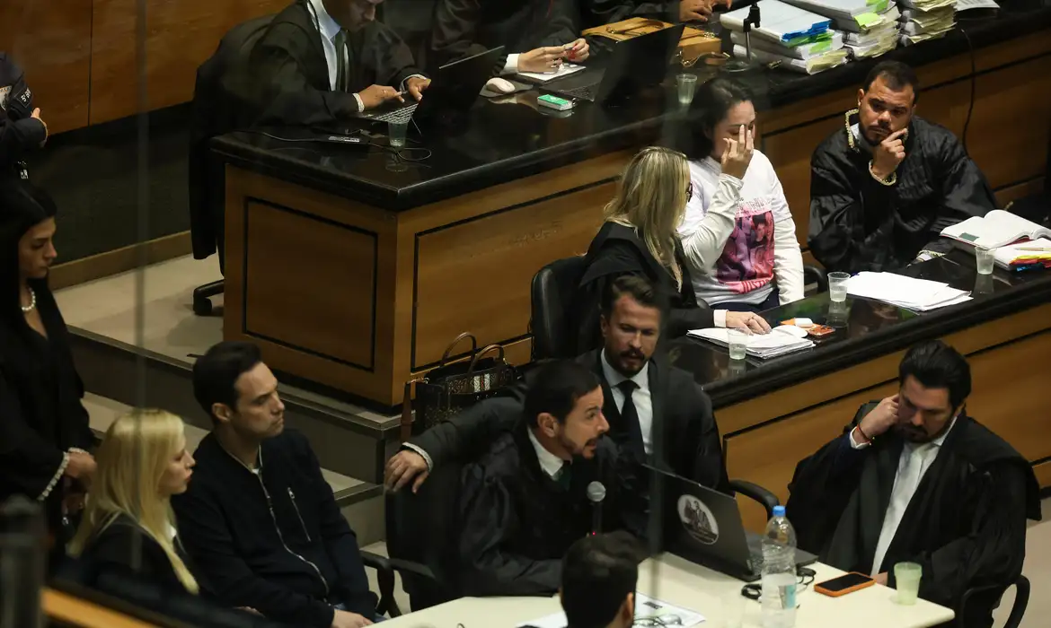 Imagem de uma babá segurando um celular com uma expressão de susto, enquanto uma policial assiste em segundo plano.