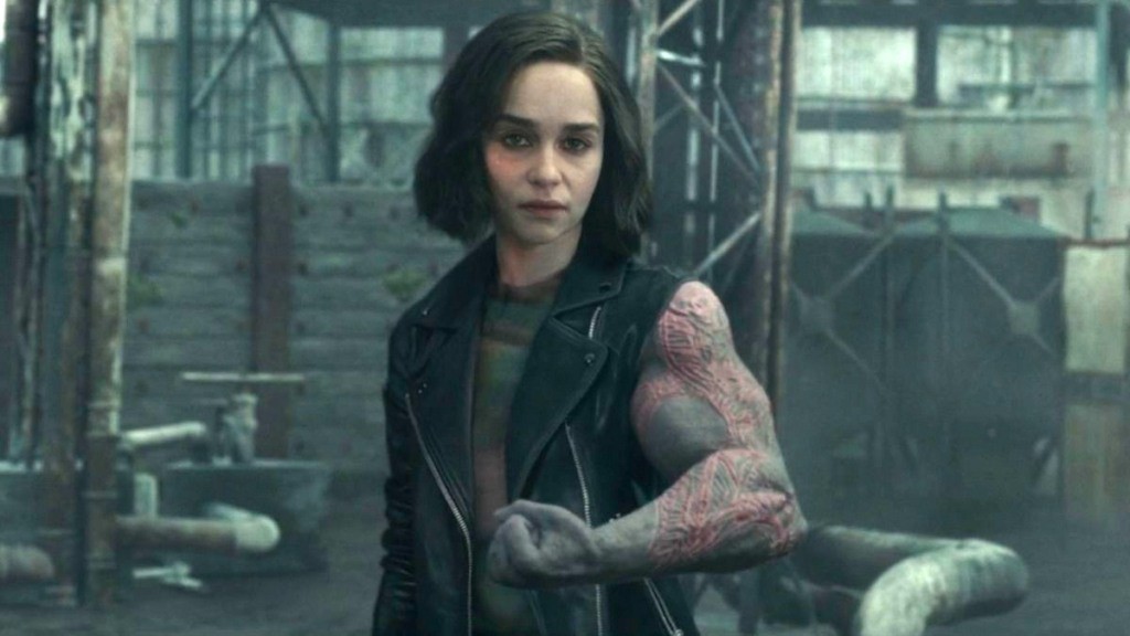 Ator Emilia Clarke sentada em frente a uma mesa de jornal, com um tom sério em seu rosto.