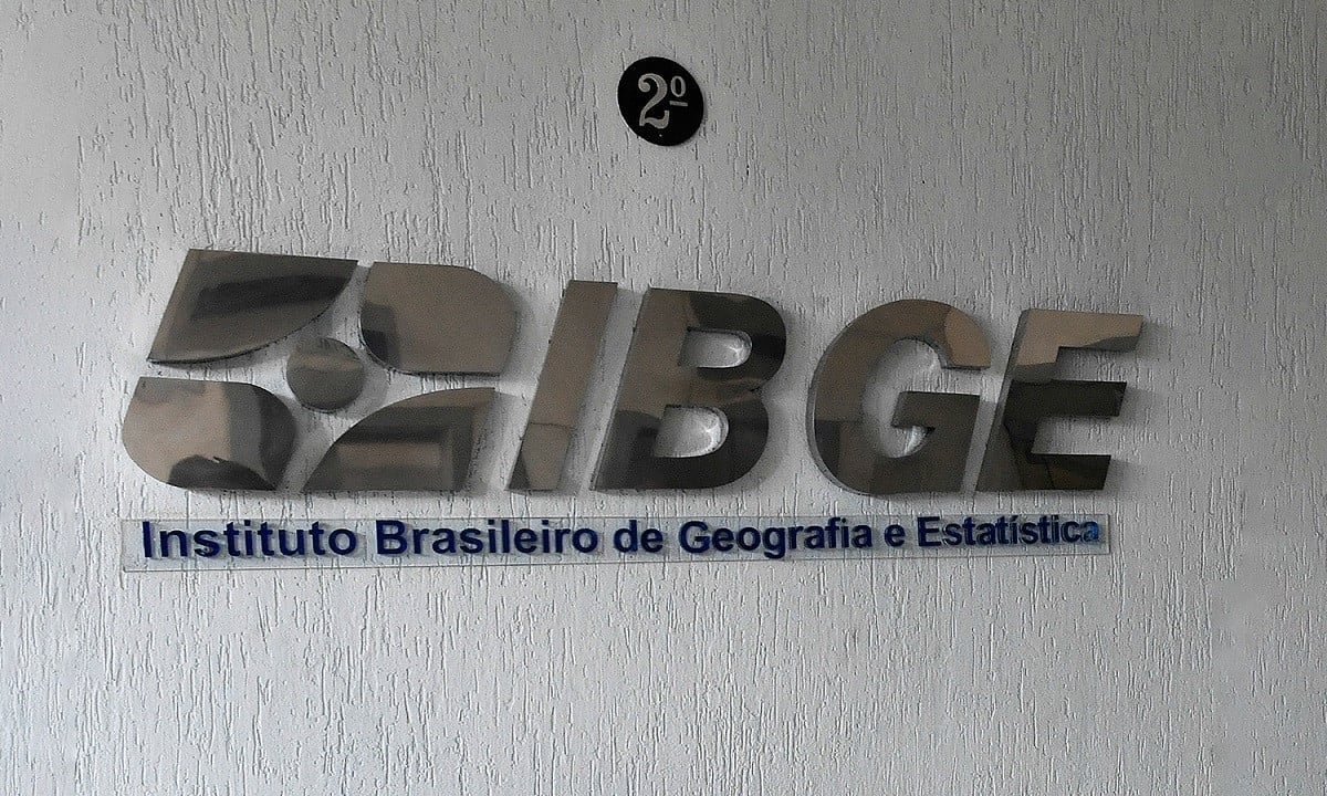 Jornalista segura papel com edital do IBGE e olha para a câmera com expressão de surpresa.