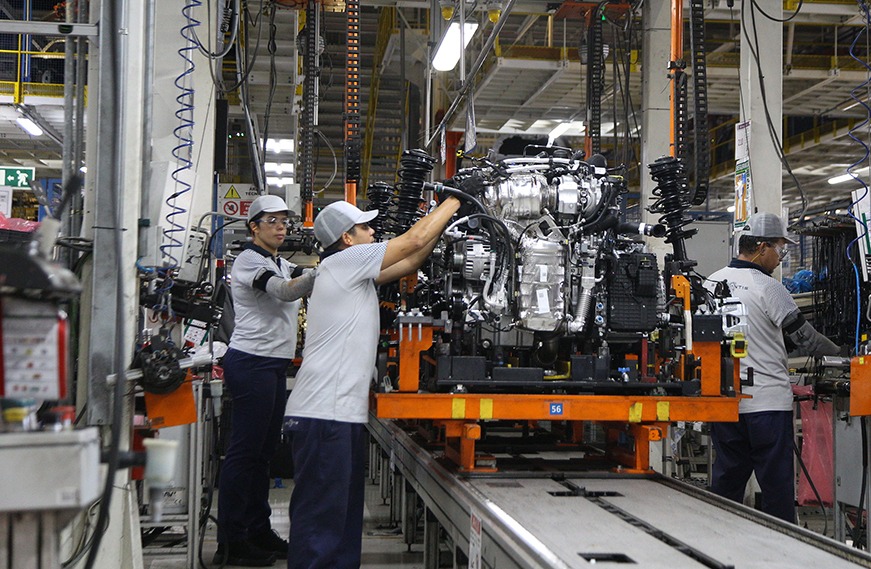Gráficos e equipamentos industriais em uma fábrica brasileira, com operários trabalhando ao fundo.