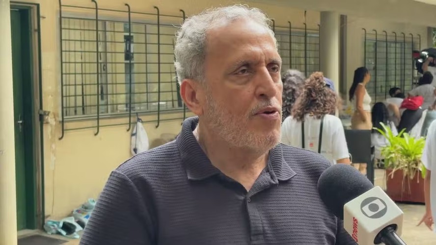 Homem idoso, com expressão triste, sentado em uma cadeira, cercado por jornalistas e câmeras, em um ambiente de noticiário.