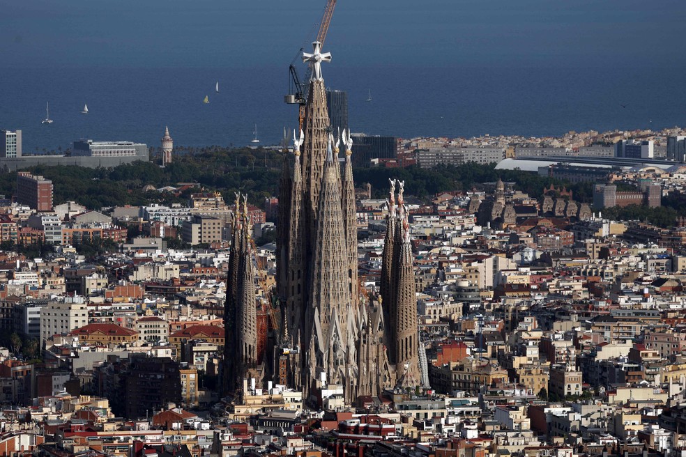 Visão aérea da Sagrada Família em construção com Papa celebrando missa ao fundo.