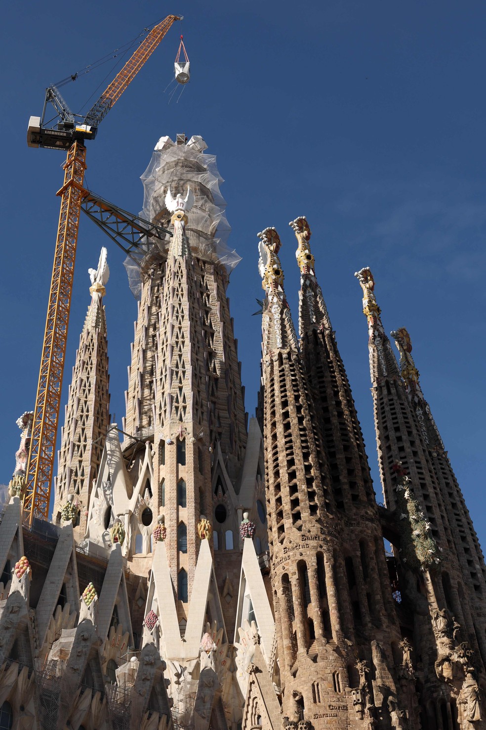 Visão aérea da Sagrada Família em construção com Papa celebrando missa ao fundo.