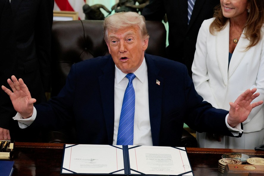 Donald Trump sorri enquanto fala sobre inflação, com fundo de notícias sobre guerra com o Irã.