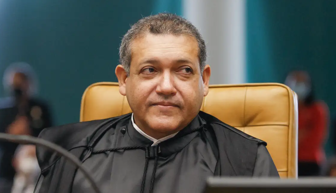 O ministro Nunes Marques, sentado à frente de uma mesa, com um rosto sério e um papel na mão.