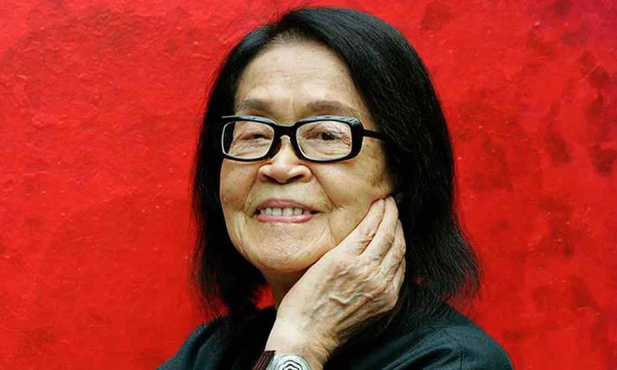 Mulher idosa sorridente, vestida de preto, recebendo homenagem em cerimônia de arte.
