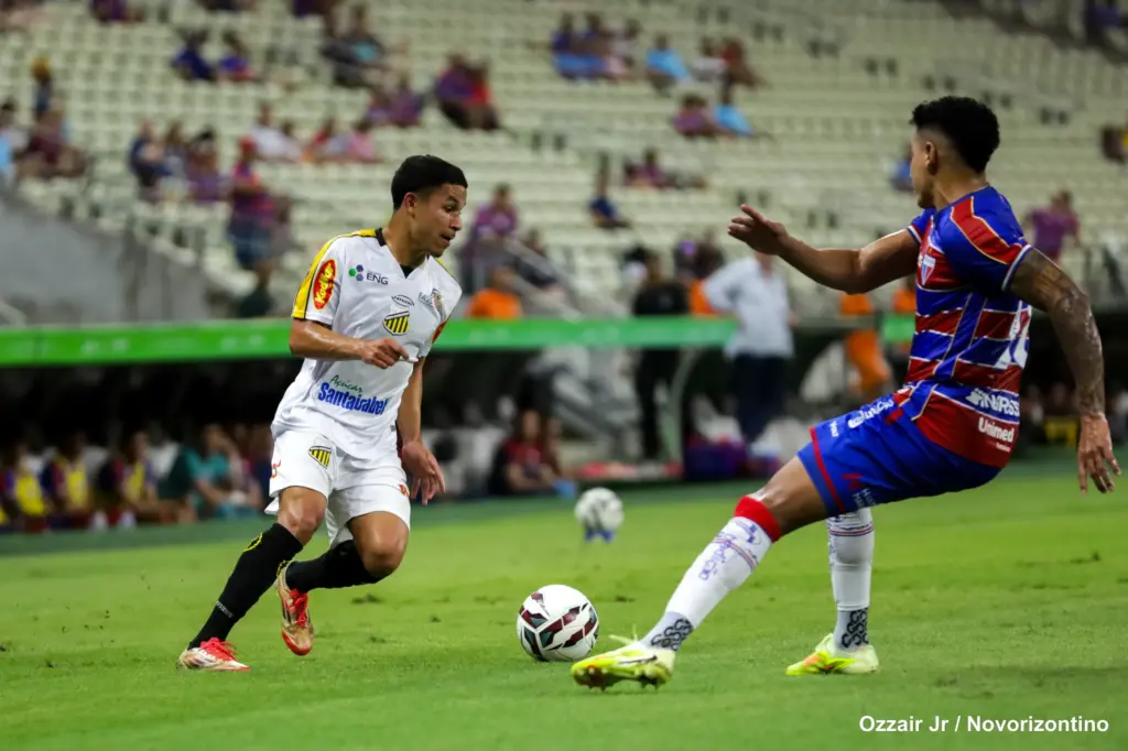 Jogador de futebol desapontado em campo, com rosto franzido e braços cruzados.