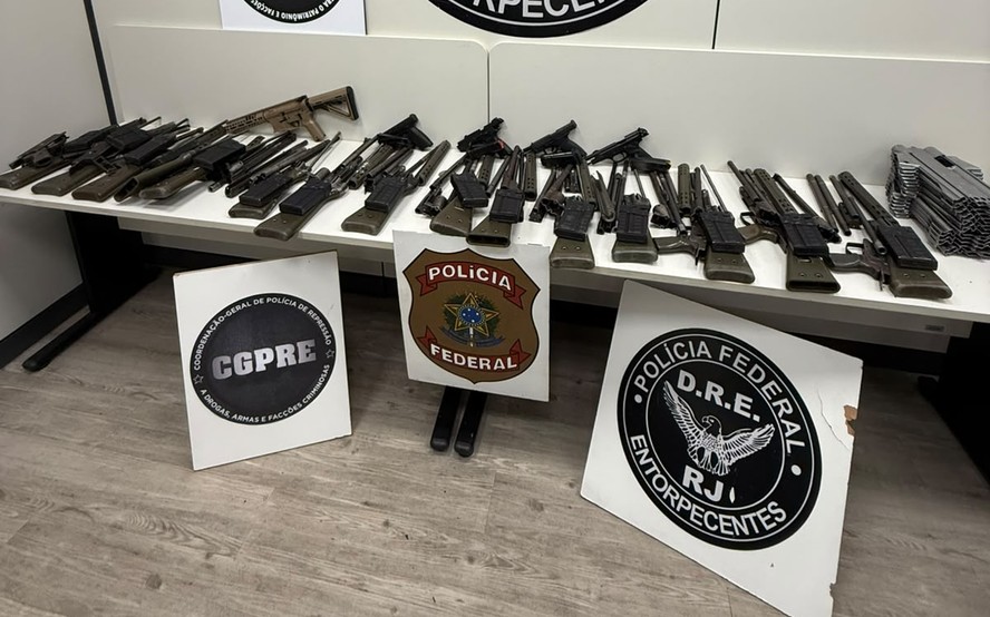 Agente do PF segura fuzis e pistolas apreendidos em operação contra facção criminosa.