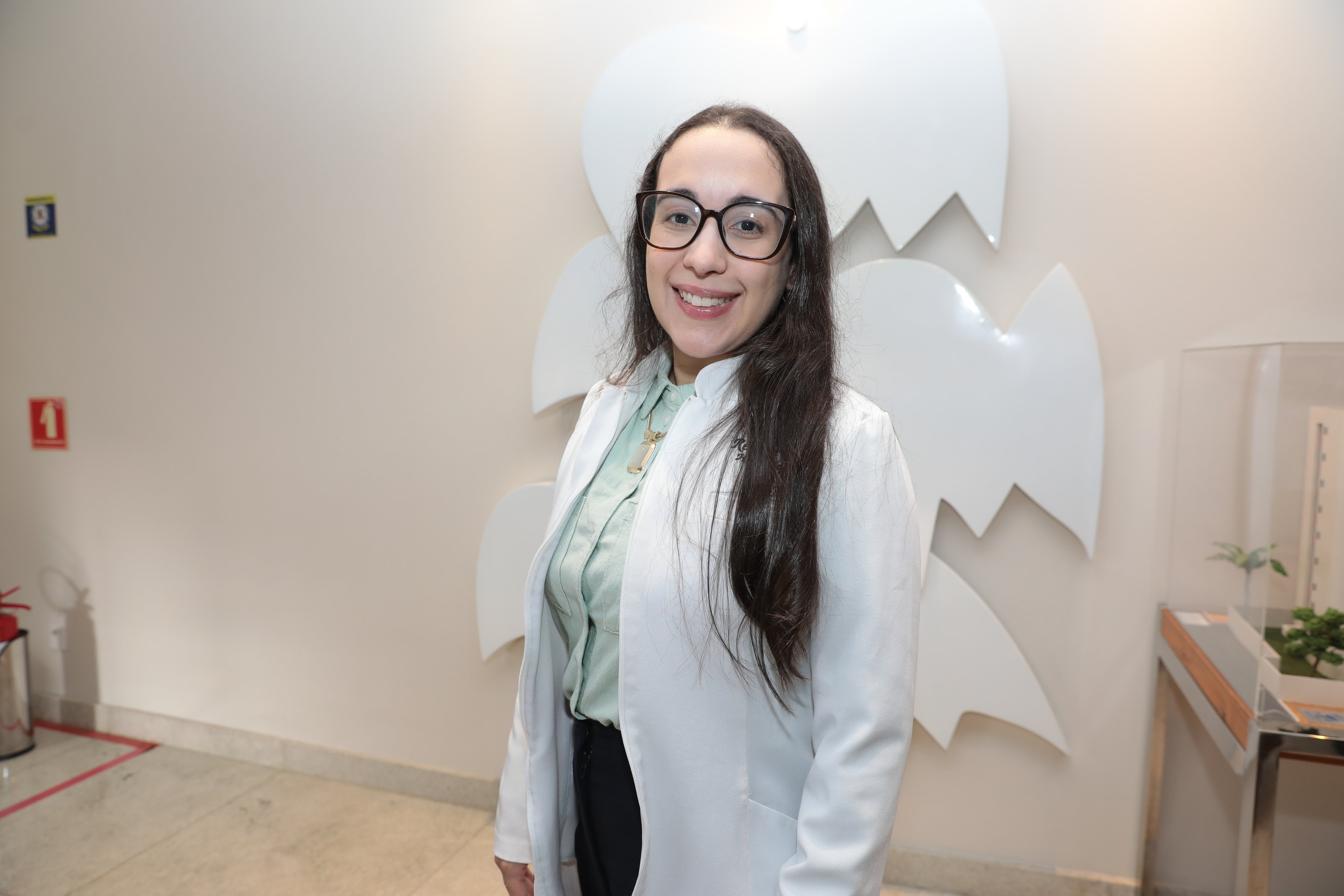 Mulher em camisa branca, com um microscópio em primeiro plano, examinando uma amostra de sangue.
