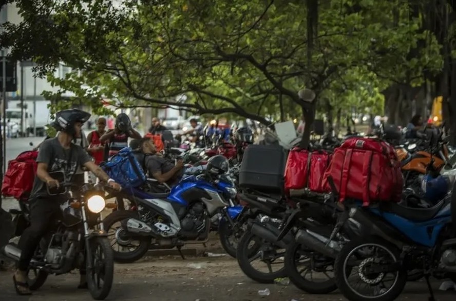 Motoristas de app e táxi aguardam em fila, com expressões preocupadas, em frente a um edifício governamental.